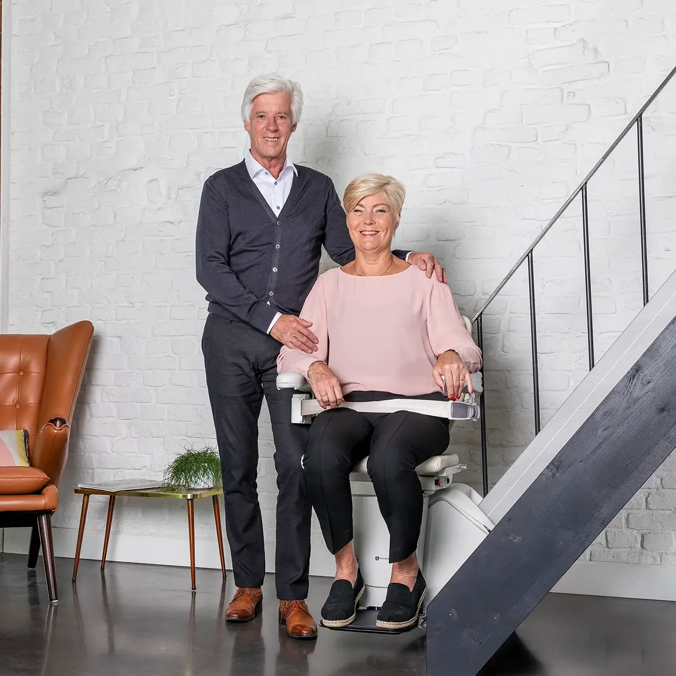 Treppenlift von der Firma Handicare. Eine Frau sitzt im Treppenlift und Ihr Mann steht neben ihr. Schleswig-Holstein Lifte führt Maschinen von Handicare.